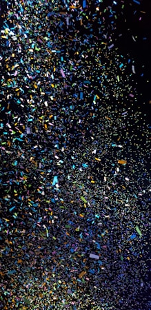 A cascade of colorful confetti falling against a dark background, creating a vibrant and dynamic scene. The confetti pieces vary in color, including shades of blue, green, pink, yellow, and orange, and they are scattered in a seemingly random pattern.