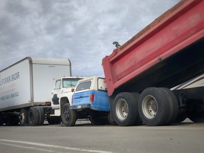 A series of trucks are parked or stationery on a road, including a white trailer with company branding, a white and blue pickup truck, and a large red dump truck with a raised bed.