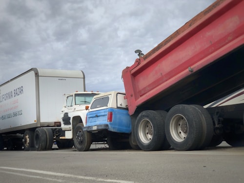 A series of trucks are parked or stationery on a road, including a white trailer with company branding, a white and blue pickup truck, and a large red dump truck with a raised bed.