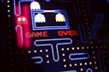 A close-up view of a classic arcade video game screen featuring familiar characters and a maze. Bright, colorful pixels form the words 'GAME OVER' in red letters in the center of the screen. Iconic ghost figures appear on the left side, while dot pathways are visible throughout the design.