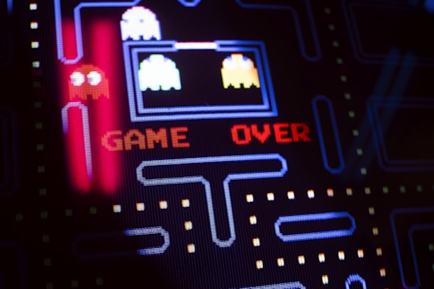 A close-up view of a classic arcade video game screen featuring familiar characters and a maze. Bright, colorful pixels form the words 'GAME OVER' in red letters in the center of the screen. Iconic ghost figures appear on the left side, while dot pathways are visible throughout the design.