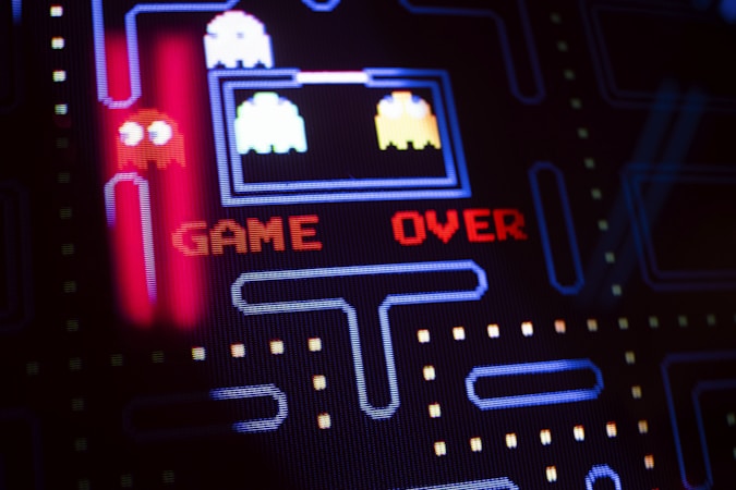 A close-up view of a classic arcade video game screen featuring familiar characters and a maze. Bright, colorful pixels form the words 'GAME OVER' in red letters in the center of the screen. Iconic ghost figures appear on the left side, while dot pathways are visible throughout the design.