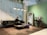 A modern living space featuring a sleek black leather sofa next to a small table with a lamp. A person is seated on the sofa, engaging with a magazine or book. The room includes a large, decorative mirror with an arch design and a wooden vanity table set against a green accent wall. The flooring is wooden, and overhead, contemporary pendant lights are hanging. Lush green plants are suspended from the ceiling.