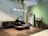 A modern living space featuring a sleek black leather sofa next to a small table with a lamp. A person is seated on the sofa, engaging with a magazine or book. The room includes a large, decorative mirror with an arch design and a wooden vanity table set against a green accent wall. The flooring is wooden, and overhead, contemporary pendant lights are hanging. Lush green plants are suspended from the ceiling.