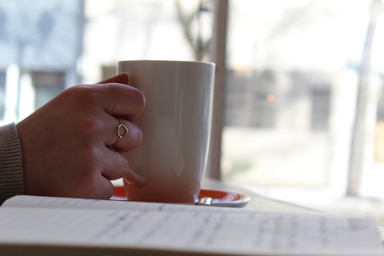 A hand holding a white mug, with a notebook in the foreground. The person is wearing a ring and a sweater. The scene appears to be set in a cozy indoor environment with soft, natural light coming from a window.