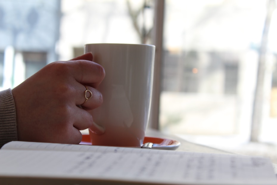A hand holding a white mug, with a notebook in the foreground. The person is wearing a ring and a sweater. The scene appears to be set in a cozy indoor environment with soft, natural light coming from a window.