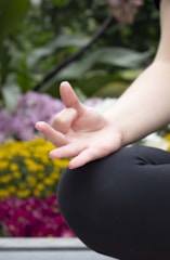 A close-up of a person's hand in a meditative position, with fingers gently touching to form a mudra. The background is filled with blurred, vibrant flowers, including yellow and purple blooms, suggesting an outdoor setting.