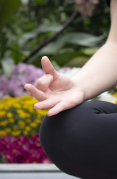 A close-up of a person's hand in a meditative position, with fingers gently touching to form a mudra. The background is filled with blurred, vibrant flowers, including yellow and purple blooms, suggesting an outdoor setting.