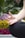 A close-up of a person's hand in a meditative position, with fingers gently touching to form a mudra. The background is filled with blurred, vibrant flowers, including yellow and purple blooms, suggesting an outdoor setting.