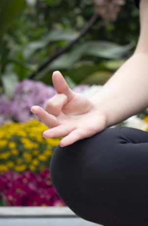 A close-up of a person's hand in a meditative position, with fingers gently touching to form a mudra. The background is filled with blurred, vibrant flowers, including yellow and purple blooms, suggesting an outdoor setting.