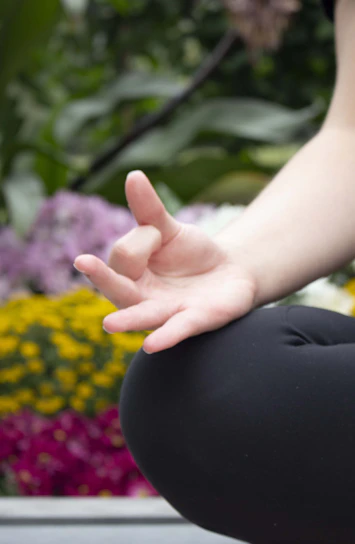 A close-up of a person's hand in a meditative position, with fingers gently touching to form a mudra. The background is filled with blurred, vibrant flowers, including yellow and purple blooms, suggesting an outdoor setting.