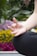 A close-up of a person's hand in a meditative position, with fingers gently touching to form a mudra. The background is filled with blurred, vibrant flowers, including yellow and purple blooms, suggesting an outdoor setting.