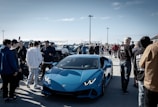 A vibrant car show on a sunny day, with a bright blue sports car prominently displayed among a crowd of people dressed in casual attire. Various vehicles are lined up in the background, and the gathering appears lively and social.