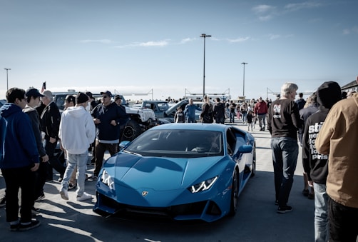 A vibrant car show on a sunny day, with a bright blue sports car prominently displayed among a crowd of people dressed in casual attire. Various vehicles are lined up in the background, and the gathering appears lively and social.