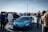 A vibrant car show on a sunny day, with a bright blue sports car prominently displayed among a crowd of people dressed in casual attire. Various vehicles are lined up in the background, and the gathering appears lively and social.