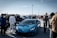 A vibrant car show on a sunny day, with a bright blue sports car prominently displayed among a crowd of people dressed in casual attire. Various vehicles are lined up in the background, and the gathering appears lively and social.