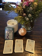 A table holds a Smith-Waite tarot deck in a tin box, three stacks of tarot cards, a lit white candle in a white holder, and a vibrant bouquet of flowers with green foliage. There is also a smooth, spherical stone with a white pattern.