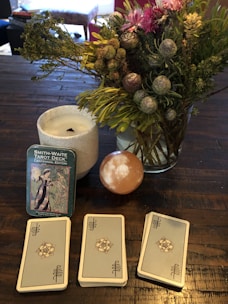 A table holds a Smith-Waite tarot deck in a tin box, three stacks of tarot cards, a lit white candle in a white holder, and a vibrant bouquet of flowers with green foliage. There is also a smooth, spherical stone with a white pattern.