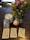 A table holds a Smith-Waite tarot deck in a tin box, three stacks of tarot cards, a lit white candle in a white holder, and a vibrant bouquet of flowers with green foliage. There is also a smooth, spherical stone with a white pattern.