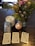 A table holds a Smith-Waite tarot deck in a tin box, three stacks of tarot cards, a lit white candle in a white holder, and a vibrant bouquet of flowers with green foliage. There is also a smooth, spherical stone with a white pattern.