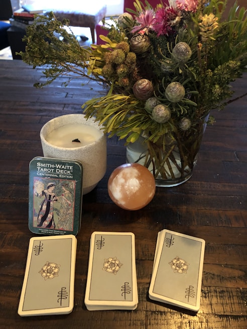 A table holds a Smith-Waite tarot deck in a tin box, three stacks of tarot cards, a lit white candle in a white holder, and a vibrant bouquet of flowers with green foliage. There is also a smooth, spherical stone with a white pattern.