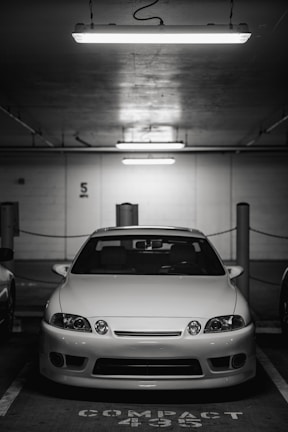 A white car is parked in an underground parking garage. It is positioned in the center of a compact parking space. Overhead, fluorescent lights illuminate the scene, casting a stark, bright light across the car's surface. The background is dimly lit and features structural details such as columns and wall markings.