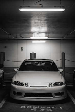A white car is parked in an underground parking garage. It is positioned in the center of a compact parking space. Overhead, fluorescent lights illuminate the scene, casting a stark, bright light across the car's surface. The background is dimly lit and features structural details such as columns and wall markings.