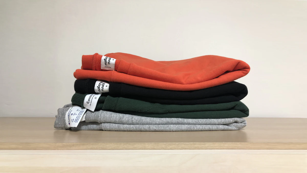 Empat sweatshirt dilipat rapi bertumpuk di rak kayu: oranye, hitam, hijau tua, dan abu-abu