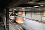 A motion blur effect captures vehicles speeding through an enclosed roadway with a gritty, urban environment. The setting includes a metal garage-like structure with corrugated doors and signage for Barbican Station. Industrial lighting casts a reflective glow on the scene.