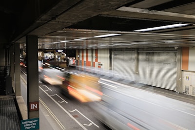 A motion blur effect captures vehicles speeding through an enclosed roadway with a gritty, urban environment. The setting includes a metal garage-like structure with corrugated doors and signage for Barbican Station. Industrial lighting casts a reflective glow on the scene.