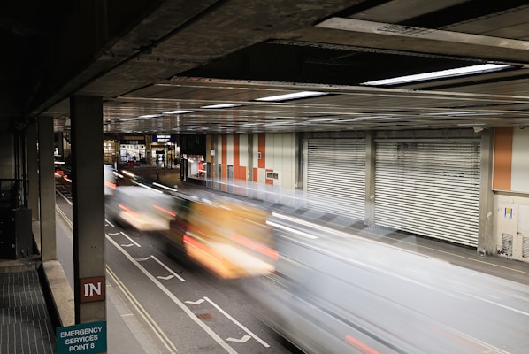 A motion blur effect captures vehicles speeding through an enclosed roadway with a gritty, urban environment. The setting includes a metal garage-like structure with corrugated doors and signage for Barbican Station. Industrial lighting casts a reflective glow on the scene.