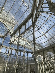 A glass and iron greenhouse with large arched windows and intricate metal framework basking in sunlight, creating reflections and shadows inside. The sky visible through the transparent ceiling is clear and blue.