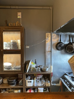 A cozy kitchen nook in a cafe features wooden cabinets filled with dishes and kitchen supplies. On top of the cabinets, there is a collection of condiments and cooking tools, adjacent to hanging frying pans on a wall. The space is organized yet bustling, giving off a homely vibe.