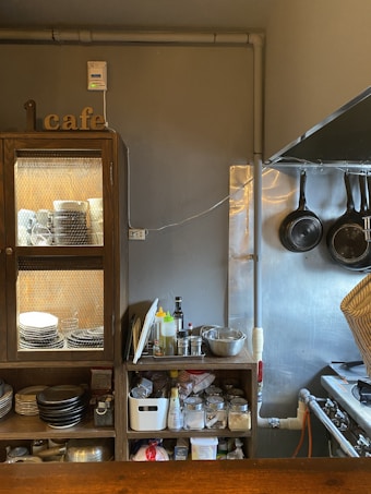 A cozy kitchen nook in a cafe features wooden cabinets filled with dishes and kitchen supplies. On top of the cabinets, there is a collection of condiments and cooking tools, adjacent to hanging frying pans on a wall. The space is organized yet bustling, giving off a homely vibe.