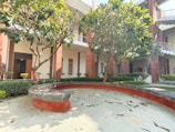 A courtyard surrounded by a two-story building with red brick walls and white accents. The area is landscaped with green bushes and trees, providing shade over a curved stone bench. Sunlight casts shadows across the tiled ground, and leaves are scattered on the surface.