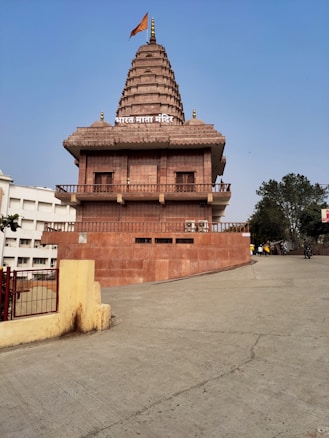 A temple with a traditional architectural style featuring a large spire. The building is constructed using reddish-brown stone. In front of the temple, there is a wide concrete pathway. Trees and other structures can be seen in the background, and there is a flag atop the temple.