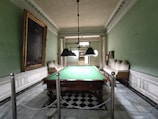 A room with a large billiards table at its center, surrounded by ornate green walls and a high ceiling with intricate moldings. Chandeliers hang above the table, providing lighting. Two couches line the walls on either side of the table, and a large framed painting is displayed on the left wall. The floor features a checkered tile pattern.