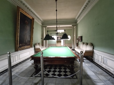 A room with a large billiards table at its center, surrounded by ornate green walls and a high ceiling with intricate moldings. Chandeliers hang above the table, providing lighting. Two couches line the walls on either side of the table, and a large framed painting is displayed on the left wall. The floor features a checkered tile pattern.