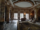 Luxurious room characterized by ornate marble columns, intricate ceiling designs with gold accents, and large arched windows. The space is furnished with elegant chairs, tables, and a sofa arranged on top of an intricately patterned rug. Soft lighting adds to the sophisticated atmosphere.