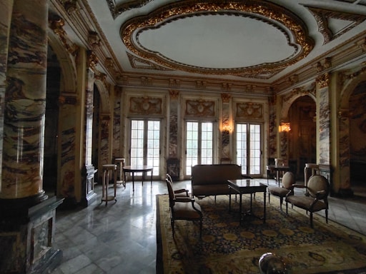Luxurious room characterized by ornate marble columns, intricate ceiling designs with gold accents, and large arched windows. The space is furnished with elegant chairs, tables, and a sofa arranged on top of an intricately patterned rug. Soft lighting adds to the sophisticated atmosphere.