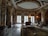 Luxurious room characterized by ornate marble columns, intricate ceiling designs with gold accents, and large arched windows. The space is furnished with elegant chairs, tables, and a sofa arranged on top of an intricately patterned rug. Soft lighting adds to the sophisticated atmosphere.
