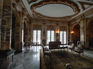 Luxurious room characterized by ornate marble columns, intricate ceiling designs with gold accents, and large arched windows. The space is furnished with elegant chairs, tables, and a sofa arranged on top of an intricately patterned rug. Soft lighting adds to the sophisticated atmosphere.