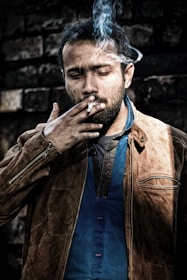 A man is wearing a brown leather jacket over a blue shirt, smoking a cigarette. Smoke is visibly swirling around his head, and he appears to be outdoors standing against a dark, textured brick wall. His expression is contemplative or introspective.