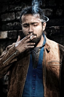 A man is wearing a brown leather jacket over a blue shirt, smoking a cigarette. Smoke is visibly swirling around his head, and he appears to be outdoors standing against a dark, textured brick wall. His expression is contemplative or introspective.