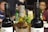Three bottles are prominently displayed on a table, with two dark wine bottles flanking a taller, amber-colored whisky bottle. In the background, there are people seated at a table, with other bottles and plates visible, suggesting a social gathering or celebration.