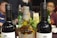 Three bottles are prominently displayed on a table, with two dark wine bottles flanking a taller, amber-colored whisky bottle. In the background, there are people seated at a table, with other bottles and plates visible, suggesting a social gathering or celebration.