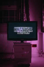A television displaying the title 'Stranger Things' sits on top of a white cabinet in a dimly lit room. The room is bathed in a pink-purple light, with patterned curtains visible in the background.