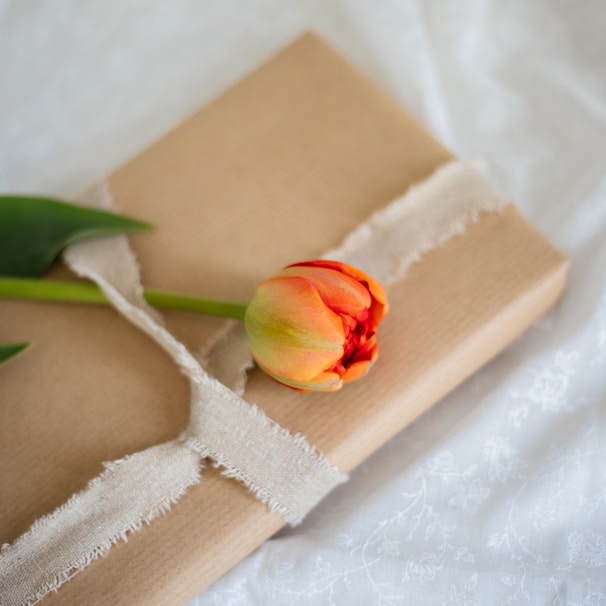 A neatly wrapped gift in simple brown paper is adorned with an elegant orange tulip. The soft frayed ribbon adds a touch of rustic charm, and the backdrop of delicate white fabric enhances the overall aesthetic.