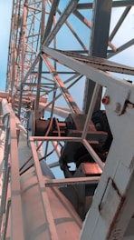 A close-up view of industrial metal structures, featuring gears, beams, and support braces. The metal appears aged and slightly rusted in some areas. The structure extends upward at an angle, filling much of the frame.