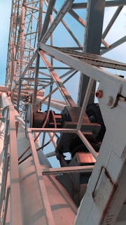 A close-up view of industrial metal structures, featuring gears, beams, and support braces. The metal appears aged and slightly rusted in some areas. The structure extends upward at an angle, filling much of the frame.